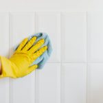 A hand in a yellow glove wiping a tiled wall with a cloth, demonstrating cleanliness.
