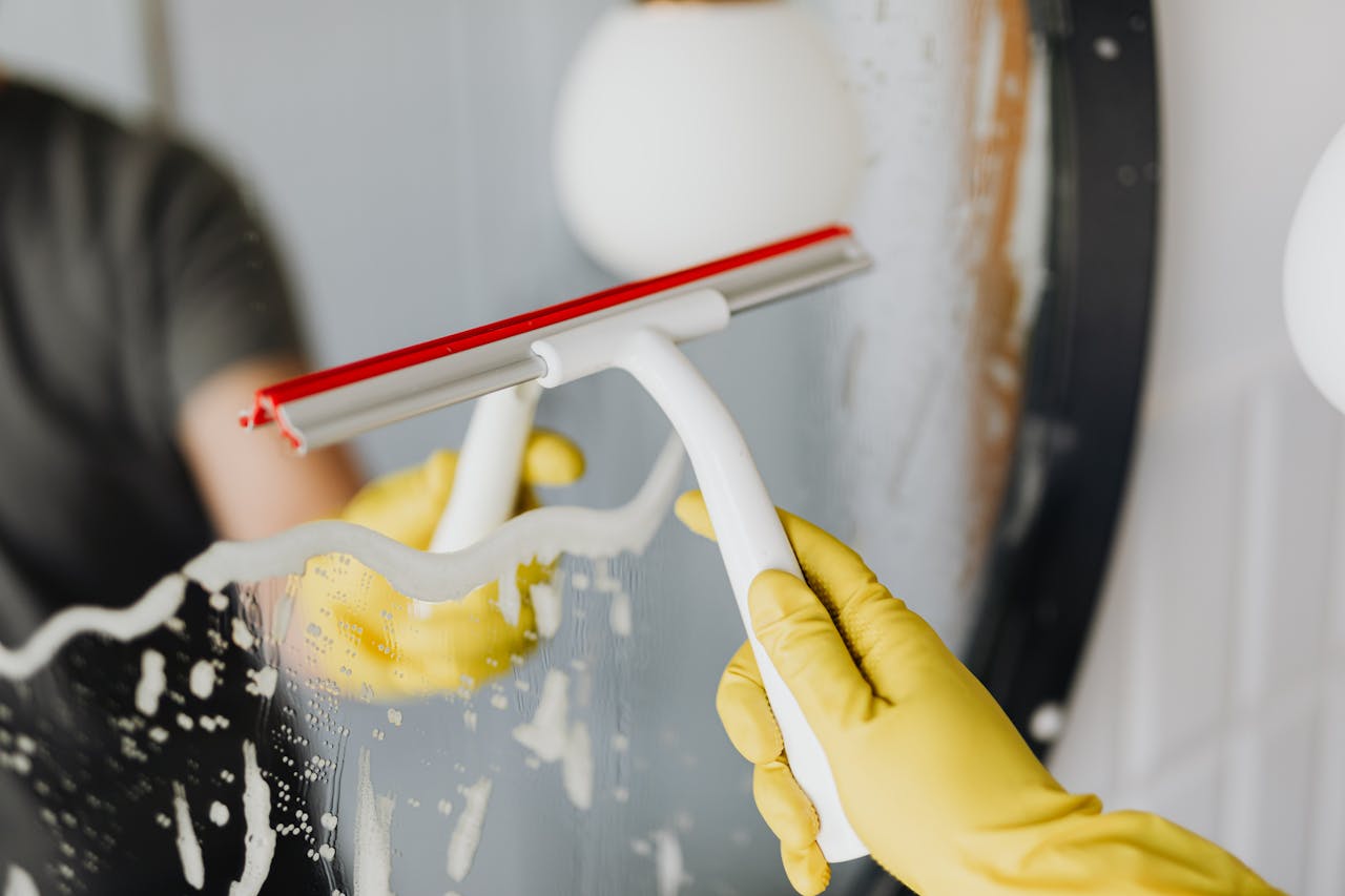 Close-up of a person cleaning a bathroom mirror with a squeegee and gloves.